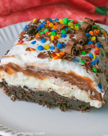 A large square slice of the Brownie Refrigerator Pudding Cake with shaved chocolate and rainbow sprinkles on the top on a white plate with a pink cloth behind it sitting on a marbled gray surface © 2026 Jane Bonacci, The Heritage Cook.