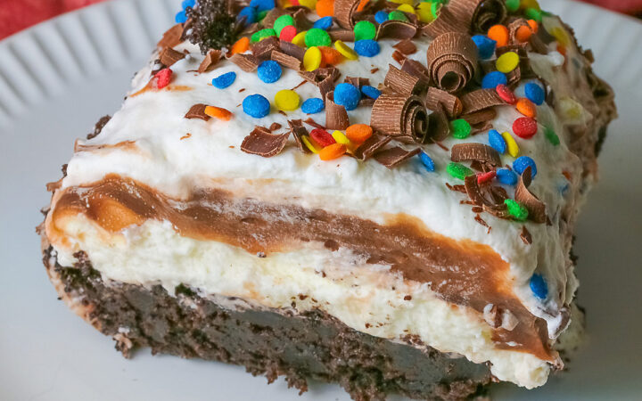 A large square slice of the Brownie Refrigerator Pudding Cake with shaved chocolate and rainbow sprinkles on the top on a white plate with a pink cloth behind it sitting on a marbled gray surface © 2026 Jane Bonacci, The Heritage Cook.