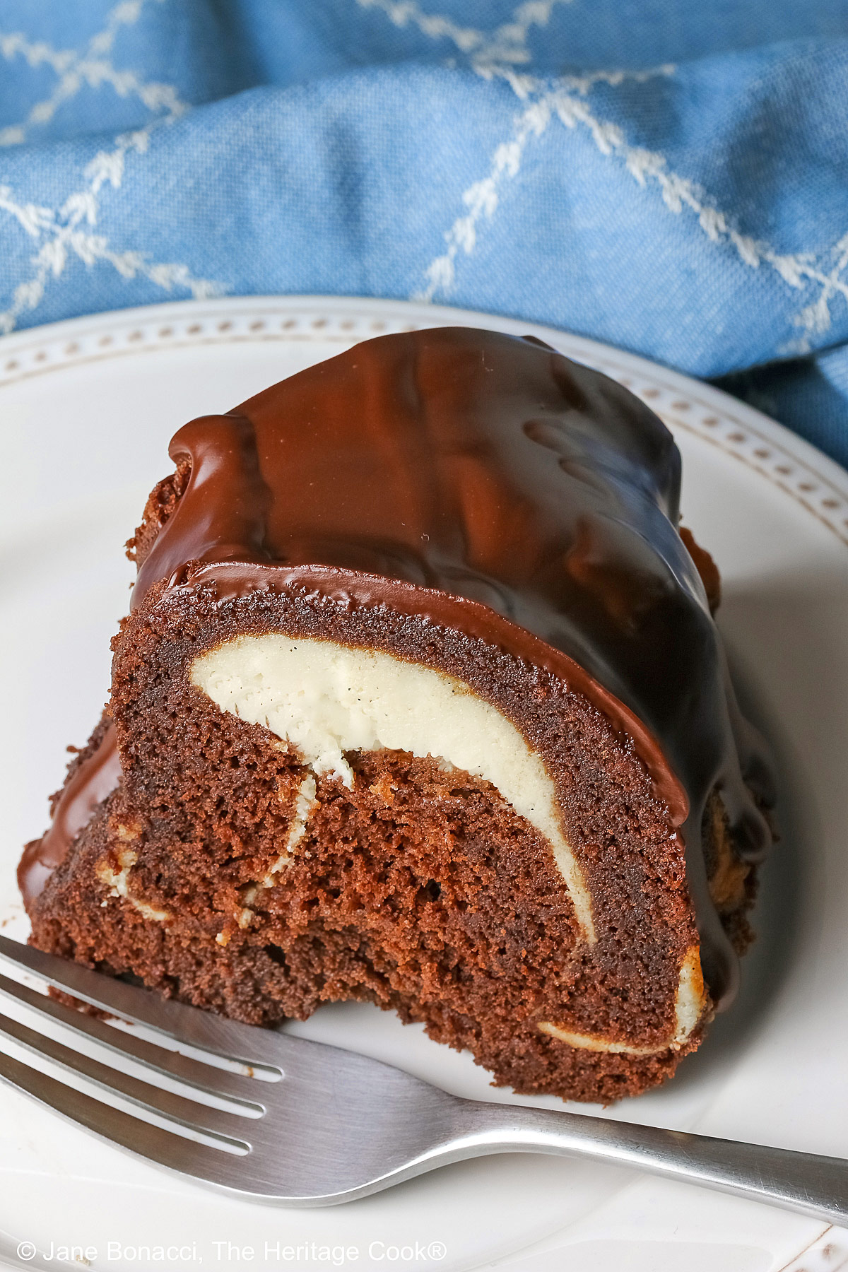 Slice of the Chocolate Bundt Cake with Cheesecake Filling on a white plate with the spiral showing and topped with thick chocolate ganache, with a blue and white cloth behind it, most with a fork on the plate © 2026 Jane Bonacci, The Heritage Cook.