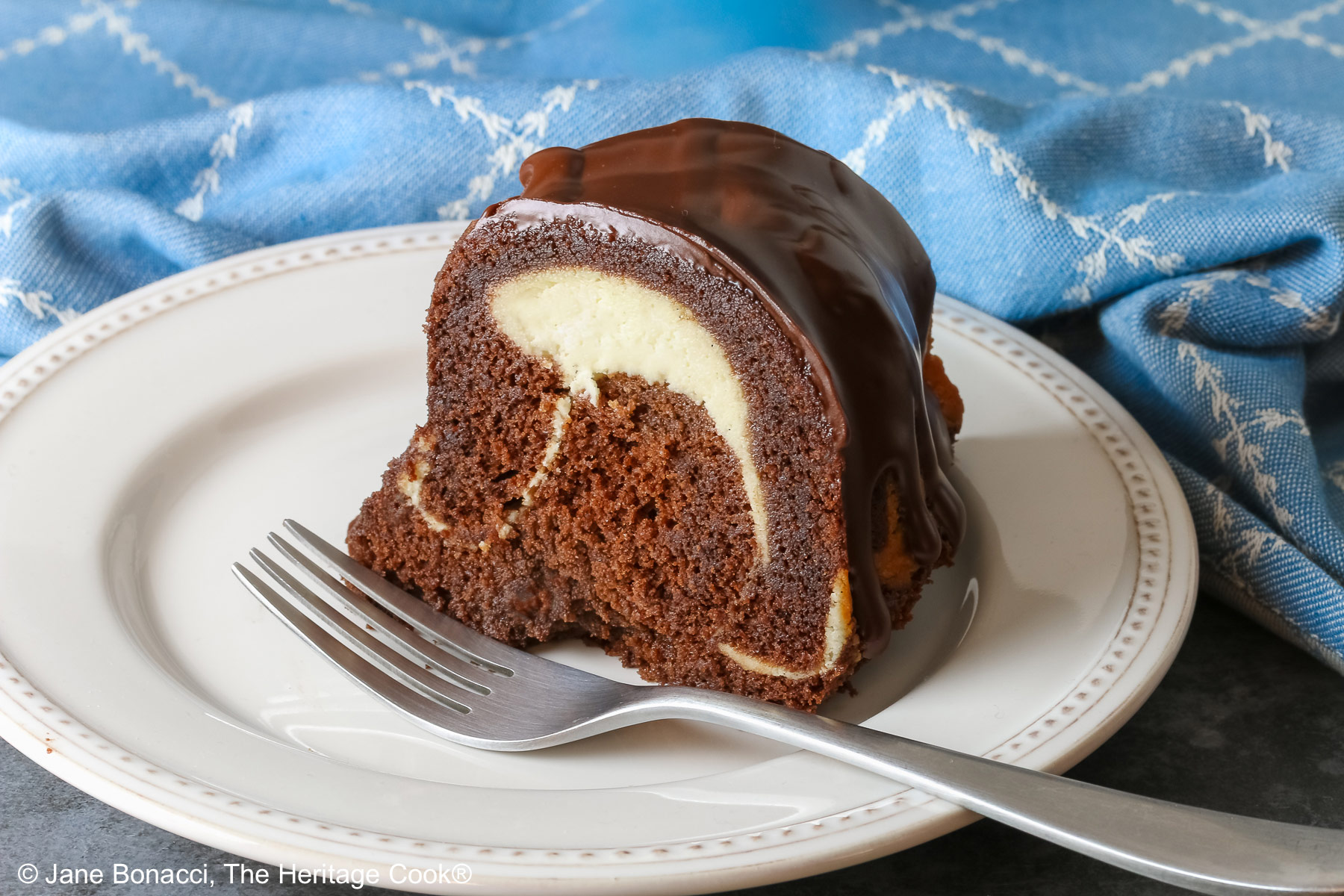Slice of the Chocolate Bundt Cake with Cheesecake Filling on a white plate with the spiral showing and topped with thick chocolate ganache, with a blue and white cloth behind it, most with a fork on the plate © 2026 Jane Bonacci, The Heritage Cook.