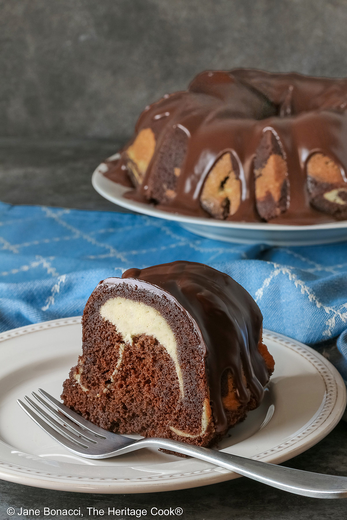Slice of the Chocolate Bundt Cake with Cheesecake Filling on a white plate with the spiral showing and topped with thick chocolate ganache, with a blue and white cloth behind it, most with a fork on the plate © 2026 Jane Bonacci, The Heritage Cook.