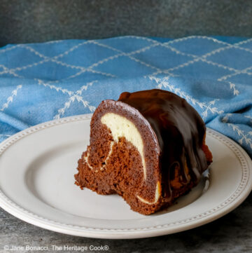 Slice of the Chocolate Bundt Cake with Cheesecake Filling on a white plate with the spiral showing and topped with thick chocolate ganache, with a blue and white cloth behind it, most with a fork on the plate © 2026 Jane Bonacci, The Heritage Cook.
