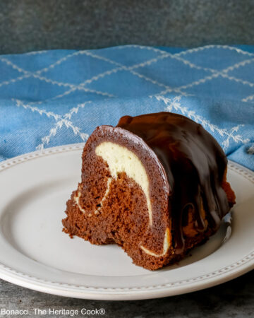 Slice of the Chocolate Bundt Cake with Cheesecake Filling on a white plate with the spiral showing and topped with thick chocolate ganache, with a blue and white cloth behind it, most with a fork on the plate © 2026 Jane Bonacci, The Heritage Cook.