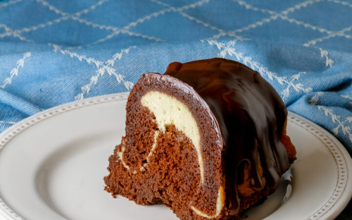 Slice of the Chocolate Bundt Cake with Cheesecake Filling on a white plate with the spiral showing and topped with thick chocolate ganache, with a blue and white cloth behind it, most with a fork on the plate © 2026 Jane Bonacci, The Heritage Cook.