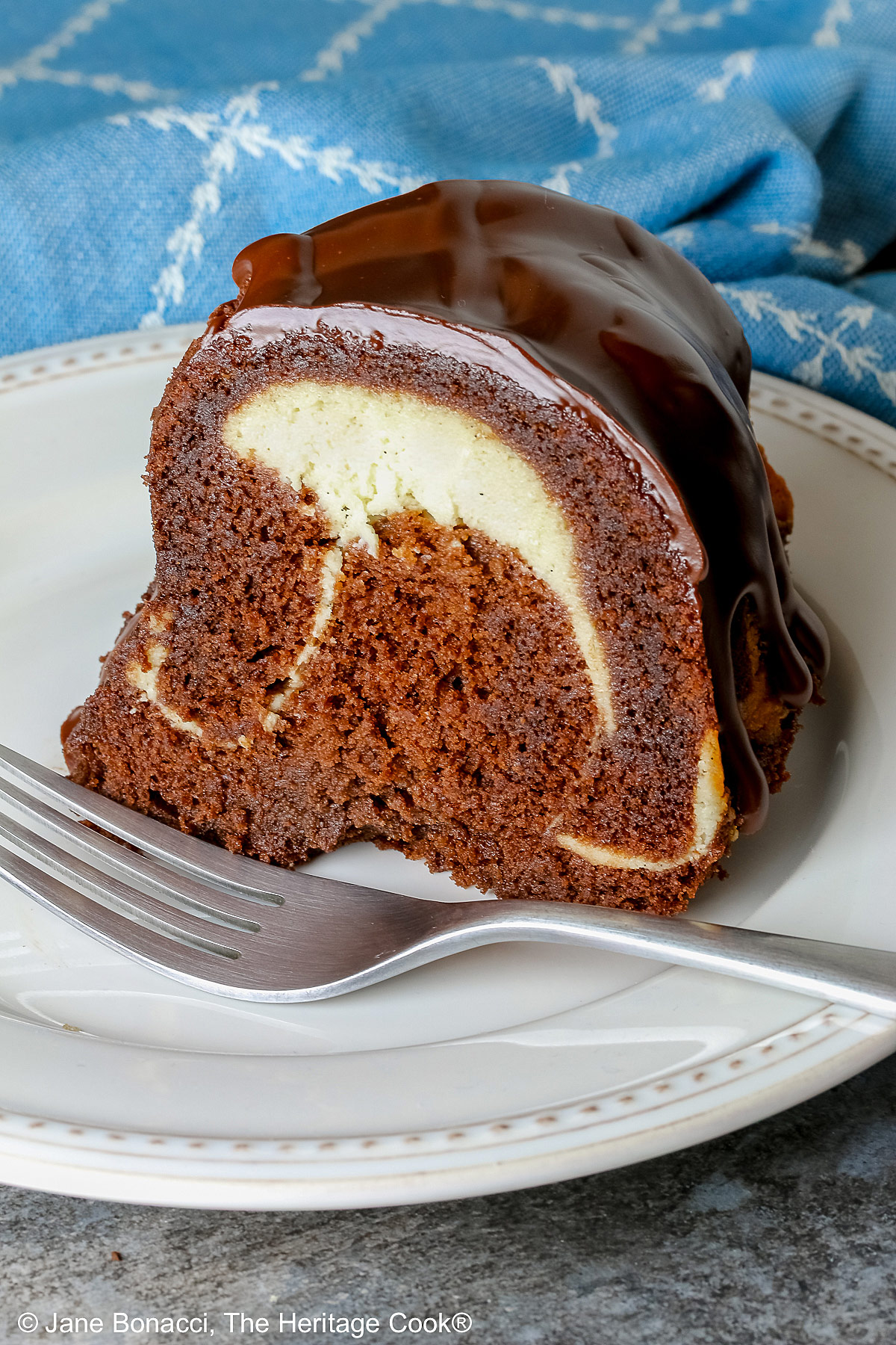 Slice of the Chocolate Bundt Cake with Cheesecake Filling on a white plate with the spiral showing and topped with thick chocolate ganache, with a blue and white cloth behind it, most with a fork on the plate © 2026 Jane Bonacci, The Heritage Cook.