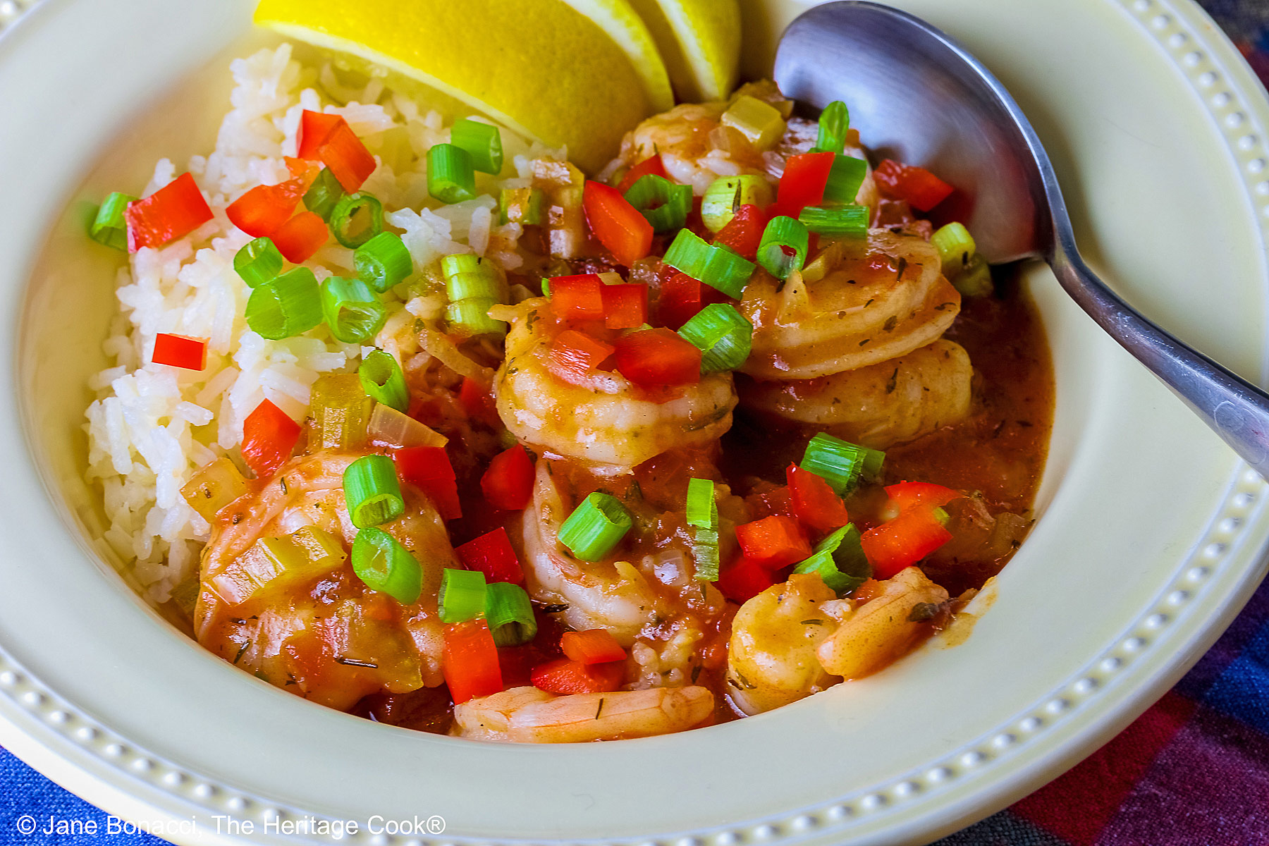 Pale yellow bowl filled with long grain rice topped with New Orleans Shrimp Creole topped with perfectly cooked shrimp, sprinkled with sliced green onions and minced red bell peppers, with 3 wedges of fresh lemon © 2026 Jane Bonacci, The Heritage Cook.