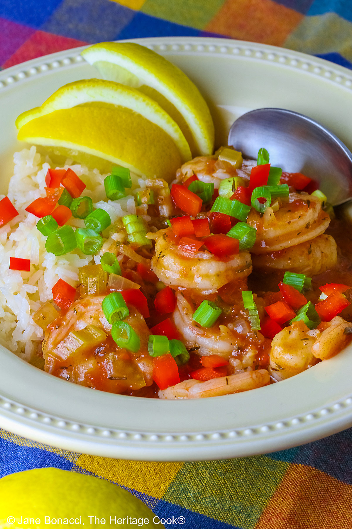Pale yellow bowl filled with long grain rice topped with New Orleans Shrimp Creole topped with perfectly cooked shrimp, sprinkled with sliced green onions and minced red bell peppers, with 3 wedges of fresh lemon © 2026 Jane Bonacci, The Heritage Cook.