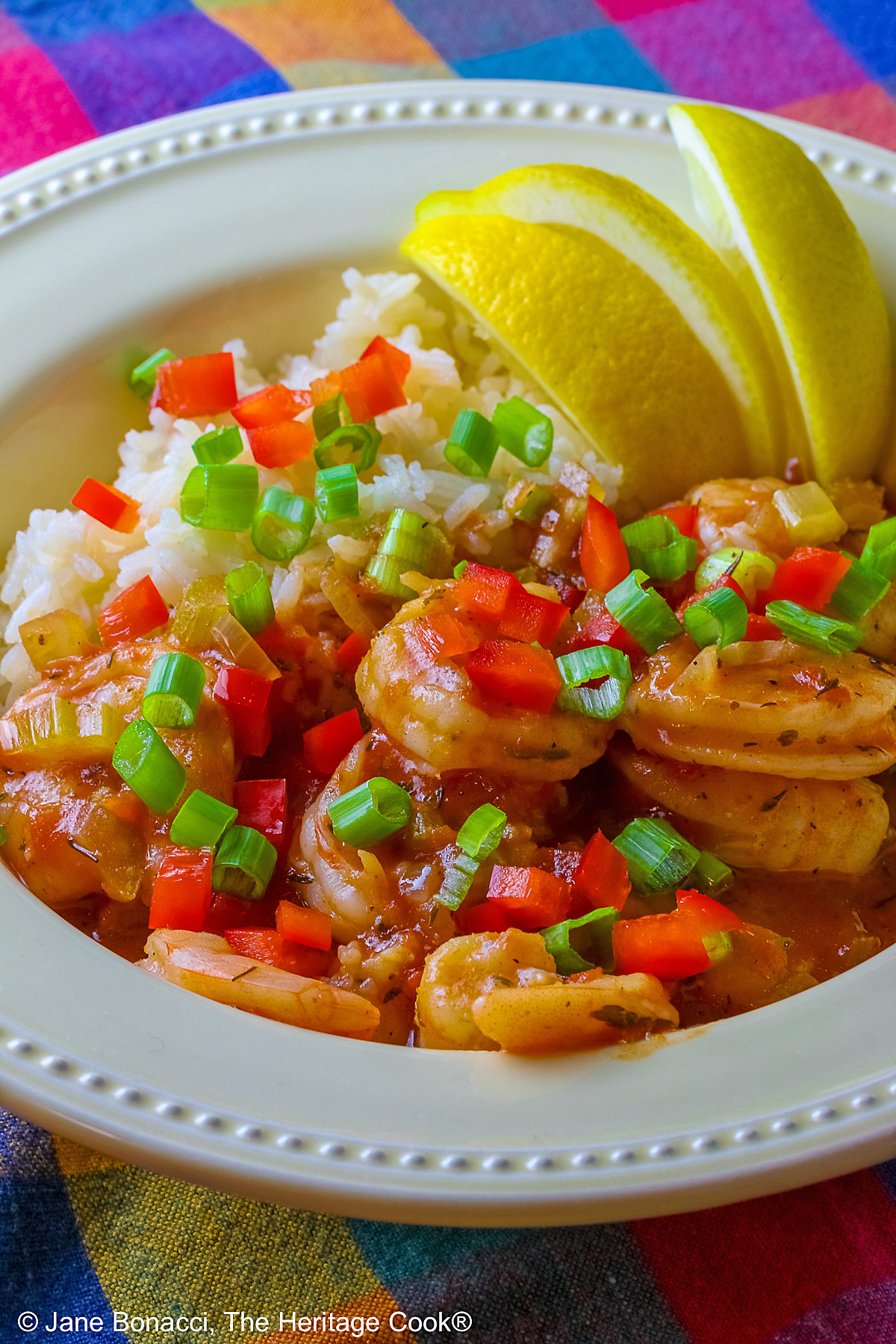 Pale yellow bowl filled with long grain rice topped with New Orleans Shrimp Creole topped with perfectly cooked shrimp, sprinkled with sliced green onions and minced red bell peppers, with 3 wedges of fresh lemon © 2026 Jane Bonacci, The Heritage Cook.