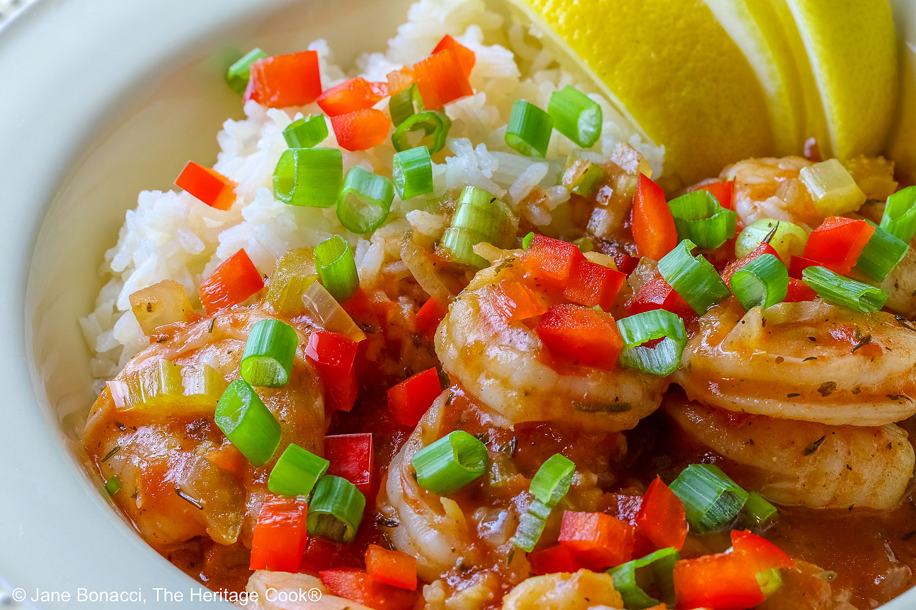 Pale yellow bowl filled with long grain rice topped with New Orleans Shrimp Creole topped with perfectly cooked shrimp, sprinkled with sliced green onions and minced red bell peppers, with 3 wedges of fresh lemon © 2026 Jane Bonacci, The Heritage Cook.