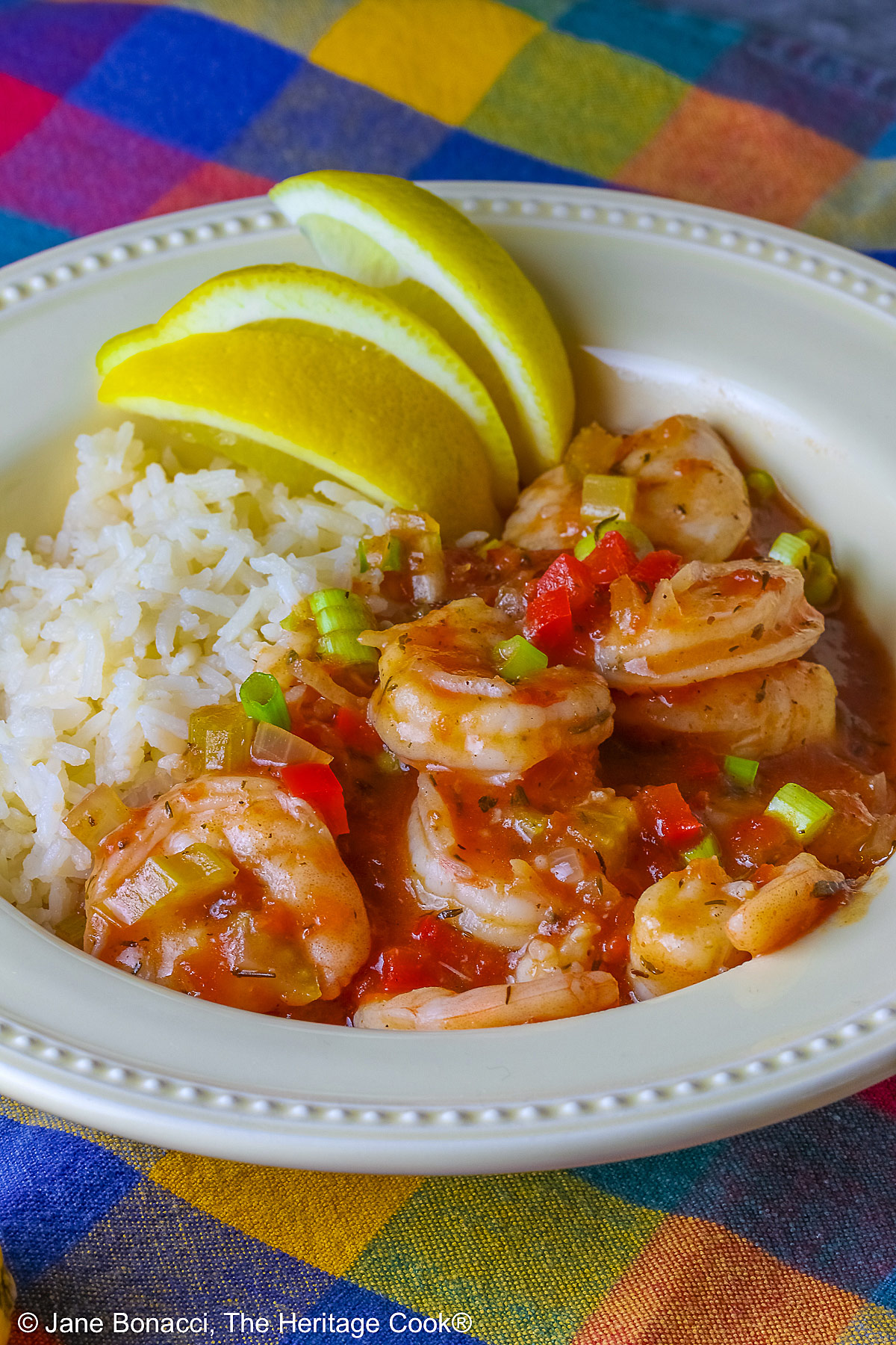 Pale yellow bowl filled with long grain rice topped with New Orleans Shrimp Creole topped with perfectly cooked shrimp, sprinkled with sliced green onions and minced red bell peppers, with 3 wedges of fresh lemon © 2026 Jane Bonacci, The Heritage Cook.