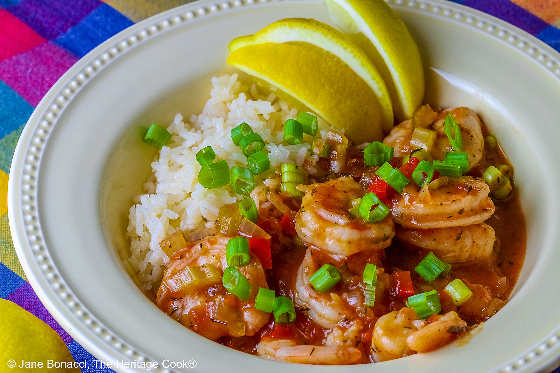 Pale yellow bowl filled with long grain rice topped with New Orleans Shrimp Creole topped with perfectly cooked shrimp, sprinkled with sliced green onions and minced red bell peppers, with 3 wedges of fresh lemon © 2026 Jane Bonacci, The Heritage Cook.