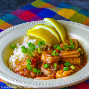 Pale yellow bowl filled with long grain rice topped with New Orleans Shrimp Creole topped with perfectly cooked shrimp, sprinkled with sliced green onions and minced red bell peppers, with 3 wedges of fresh lemon © 2026 Jane Bonacci, The Heritage Cook.