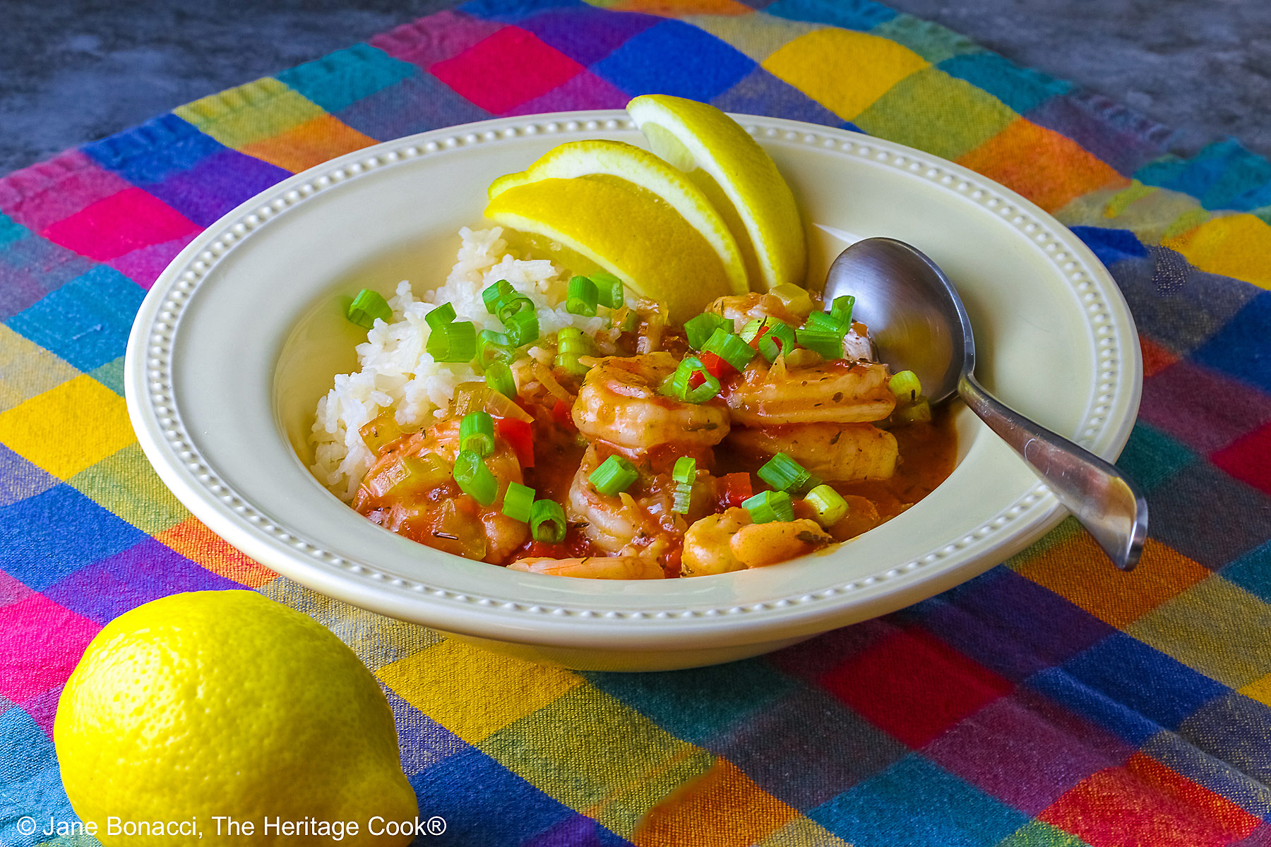 Pale yellow bowl filled with long grain rice topped with New Orleans Shrimp Creole topped with perfectly cooked shrimp, sprinkled with sliced green onions and minced red bell peppers, with 3 wedges of fresh lemon © 2026 Jane Bonacci, The Heritage Cook.