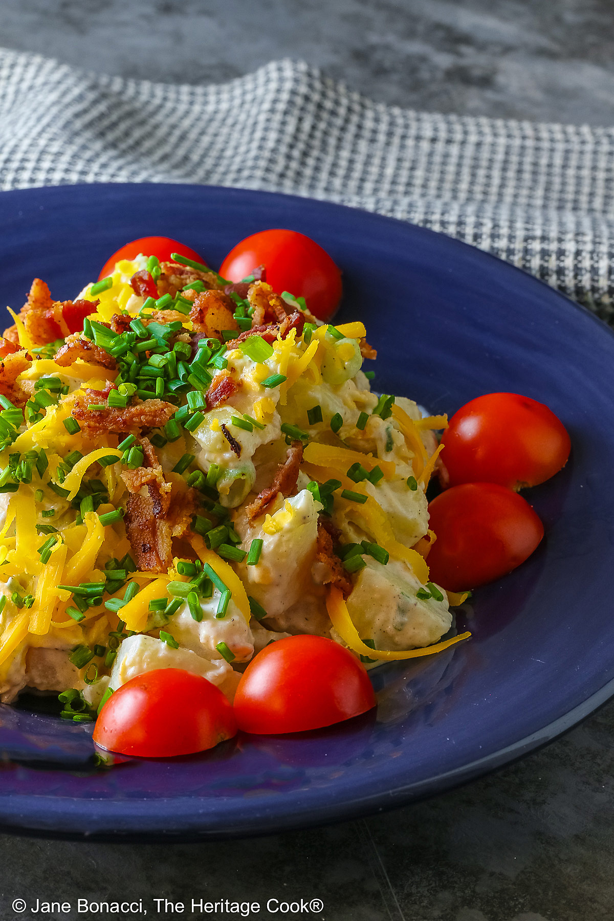 A large serving of Sassy Loaded Baked Potato Salad in a dark blue bowl with a wide rim, and a grey and black checked cloth behind, topped with shredded cheddar cheese, bacon bits, chives, and bright red cherry tomatoes © 2026 Jane Bonacci, The Heritage Cook.