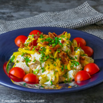 A large serving of Sassy Loaded Baked Potato Salad in a dark blue bowl with a wide rim, and a grey and black checked cloth behind, topped with shredded cheddar cheese, bacon bits, chives, and bright red cherry tomatoes © 2026 Jane Bonacci, The Heritage Cook.