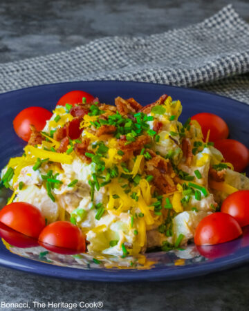A large serving of Sassy Loaded Baked Potato Salad in a dark blue bowl with a wide rim, and a grey and black checked cloth behind, topped with shredded cheddar cheese, bacon bits, chives, and bright red cherry tomatoes © 2026 Jane Bonacci, The Heritage Cook.