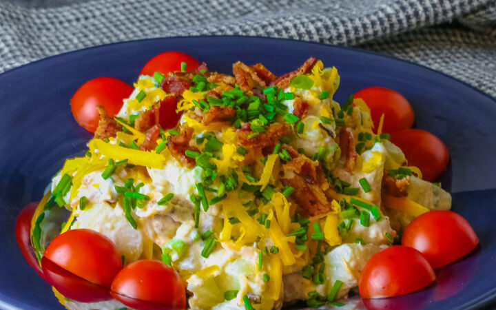 A large serving of Sassy Loaded Baked Potato Salad in a dark blue bowl with a wide rim, and a grey and black checked cloth behind, topped with shredded cheddar cheese, bacon bits, chives, and bright red cherry tomatoes © 2026 Jane Bonacci, The Heritage Cook.
