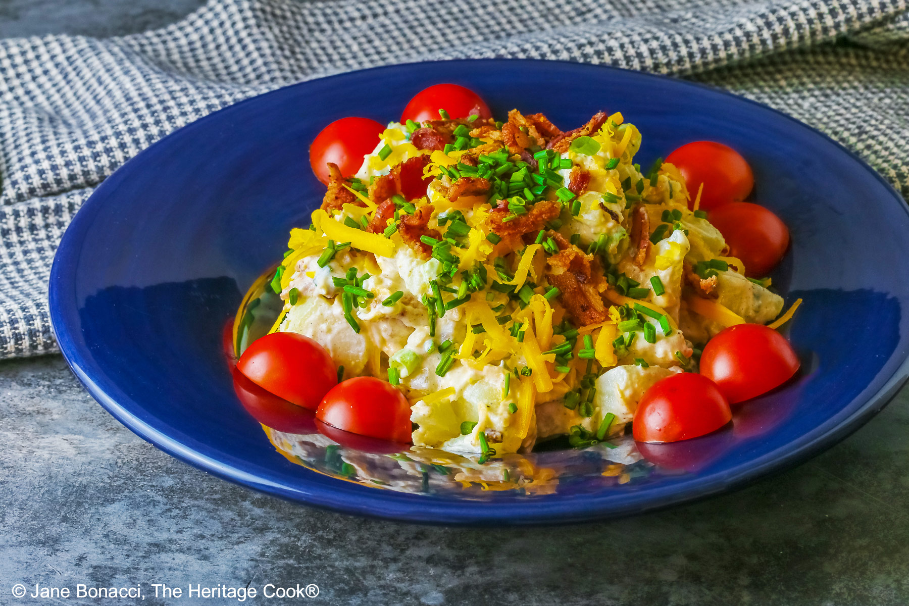 A large serving of Sassy Loaded Baked Potato Salad in a dark blue bowl with a wide rim, and a grey and black checked cloth behind, topped with shredded cheddar cheese, bacon bits, chives, and bright red cherry tomatoes © 2026 Jane Bonacci, The Heritage Cook.