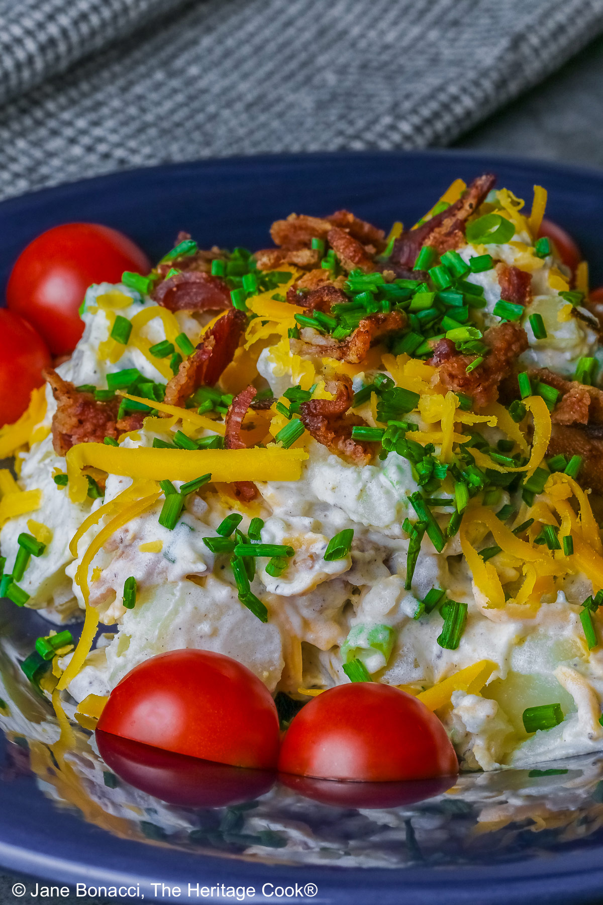 A large serving of Sassy Loaded Baked Potato Salad in a dark blue bowl with a wide rim, and a grey and black checked cloth behind, topped with shredded cheddar cheese, bacon bits, chives, and bright red cherry tomatoes © 2026 Jane Bonacci, The Heritage Cook.
