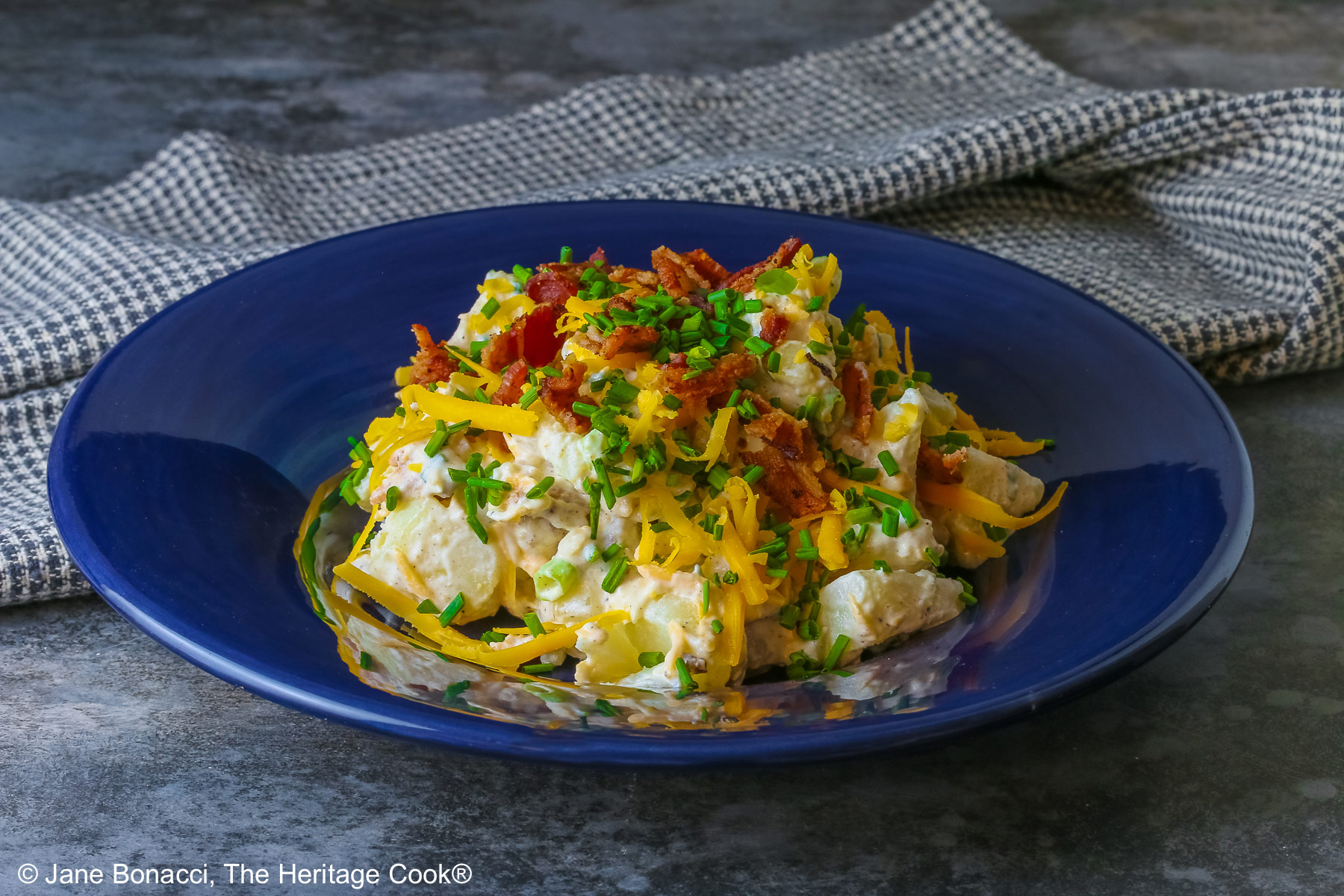 A large serving of Sassy Loaded Baked Potato Salad in a dark blue bowl with a wide rim, and a grey and black checked cloth behind, topped with shredded cheddar cheese, bacon bits, chives, and bright red cherry tomatoes © 2026 Jane Bonacci, The Heritage Cook.