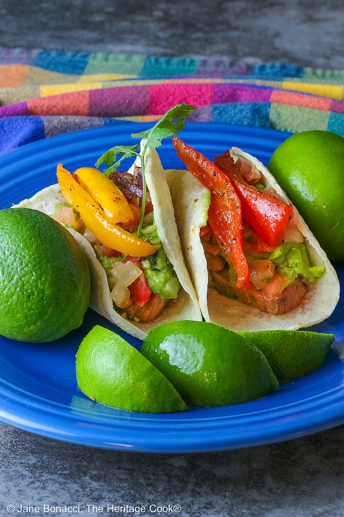 Two One-Pan Steak Fajitas tacos in corn tortillas on a dark blue plate, topped with guacamole, pico de gallo salsa, roasted onions, and bell pepper strips, surrounded by whole limes and lime wedges, with a multi-colored cloth behind and gray background © 2026 Jane Bonacci, The Heritage Cook.