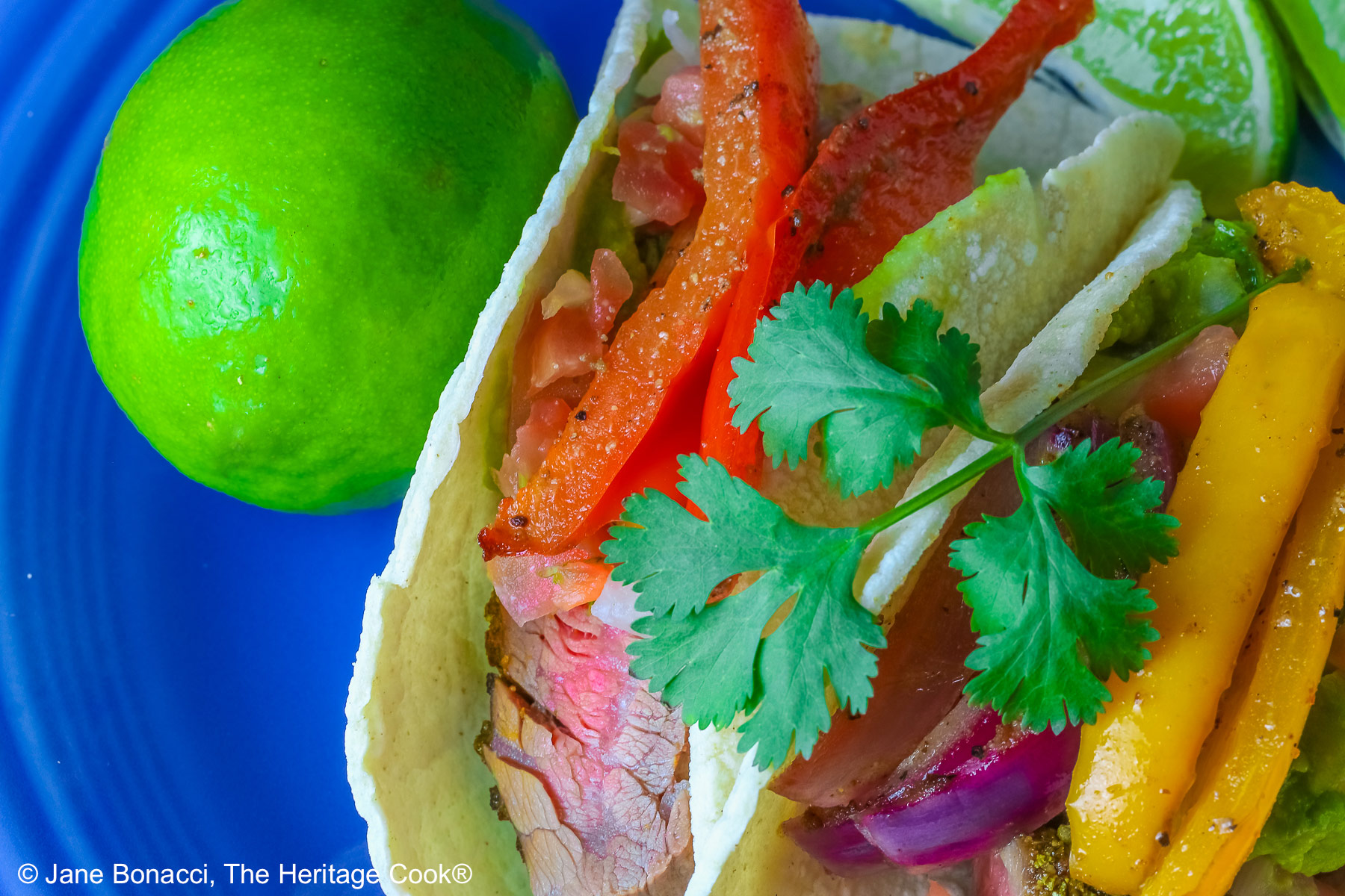 Two One-Pan Steak Fajitas tacos in corn tortillas on a dark blue plate, topped with guacamole, pico de gallo salsa, roasted onions, and bell pepper strips, surrounded by whole limes and lime wedges, with a multi-colored cloth behind and gray background © 2026 Jane Bonacci, The Heritage Cook.