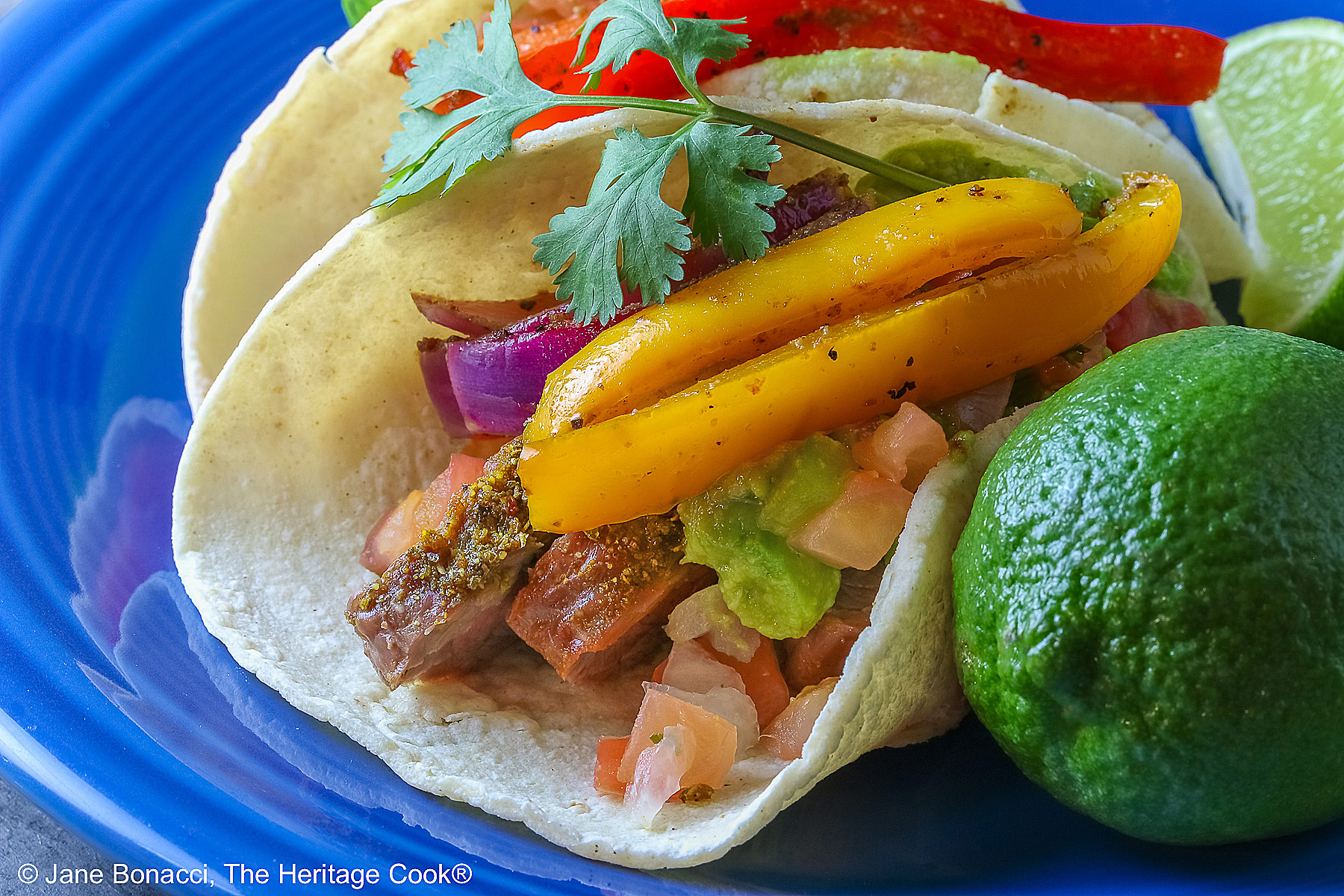 Two One-Pan Steak Fajitas tacos in corn tortillas on a dark blue plate, topped with guacamole, pico de gallo salsa, roasted onions, and bell pepper strips, surrounded by whole limes and lime wedges, with a multi-colored cloth behind and gray background © 2026 Jane Bonacci, The Heritage Cook.