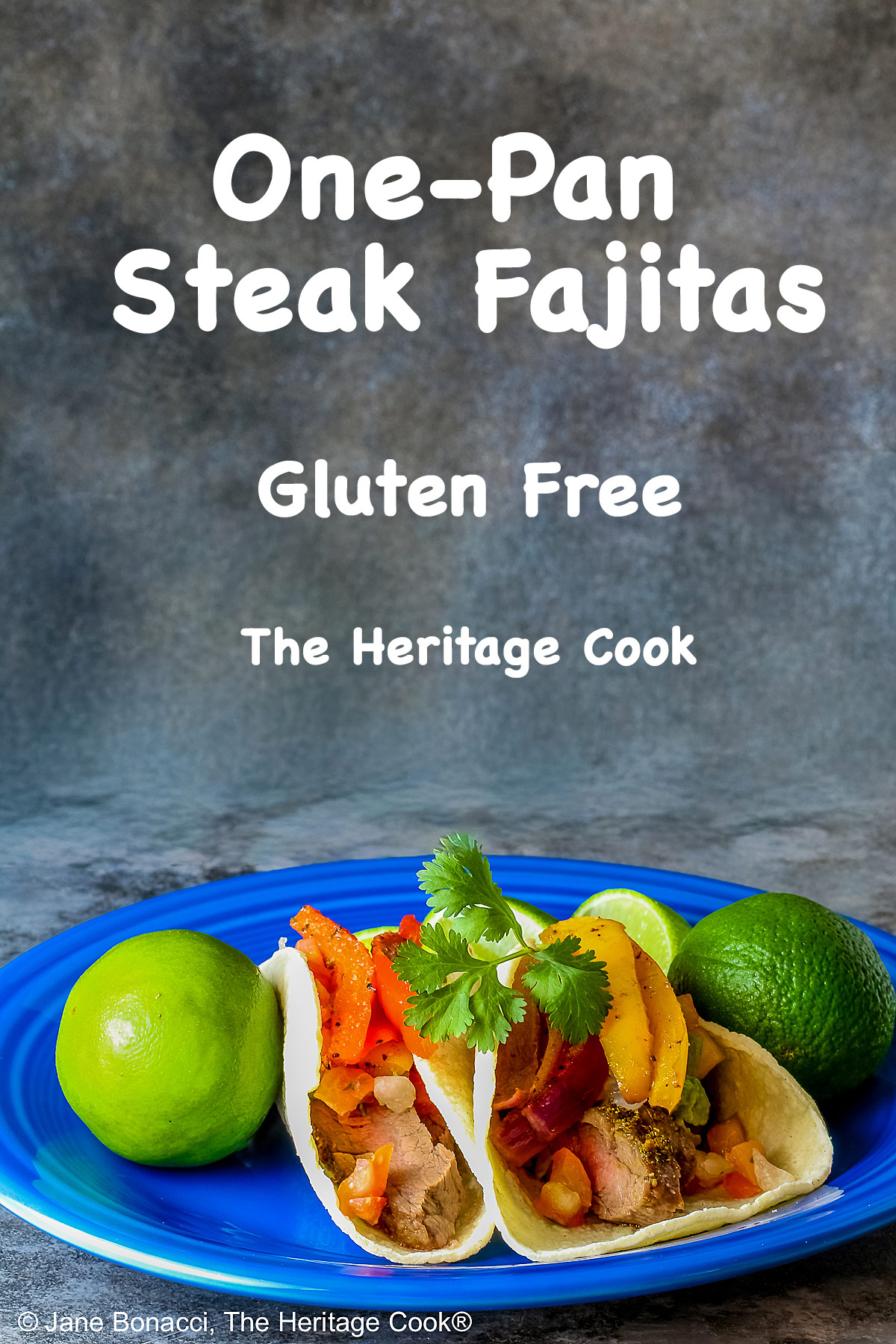 Two One-Pan Steak Fajitas tacos in corn tortillas on a dark blue plate, topped with guacamole, pico de gallo salsa, roasted onions, and bell pepper strips, surrounded by whole limes and lime wedges, with a multi-colored cloth behind and gray background © 2026 Jane Bonacci, The Heritage Cook.