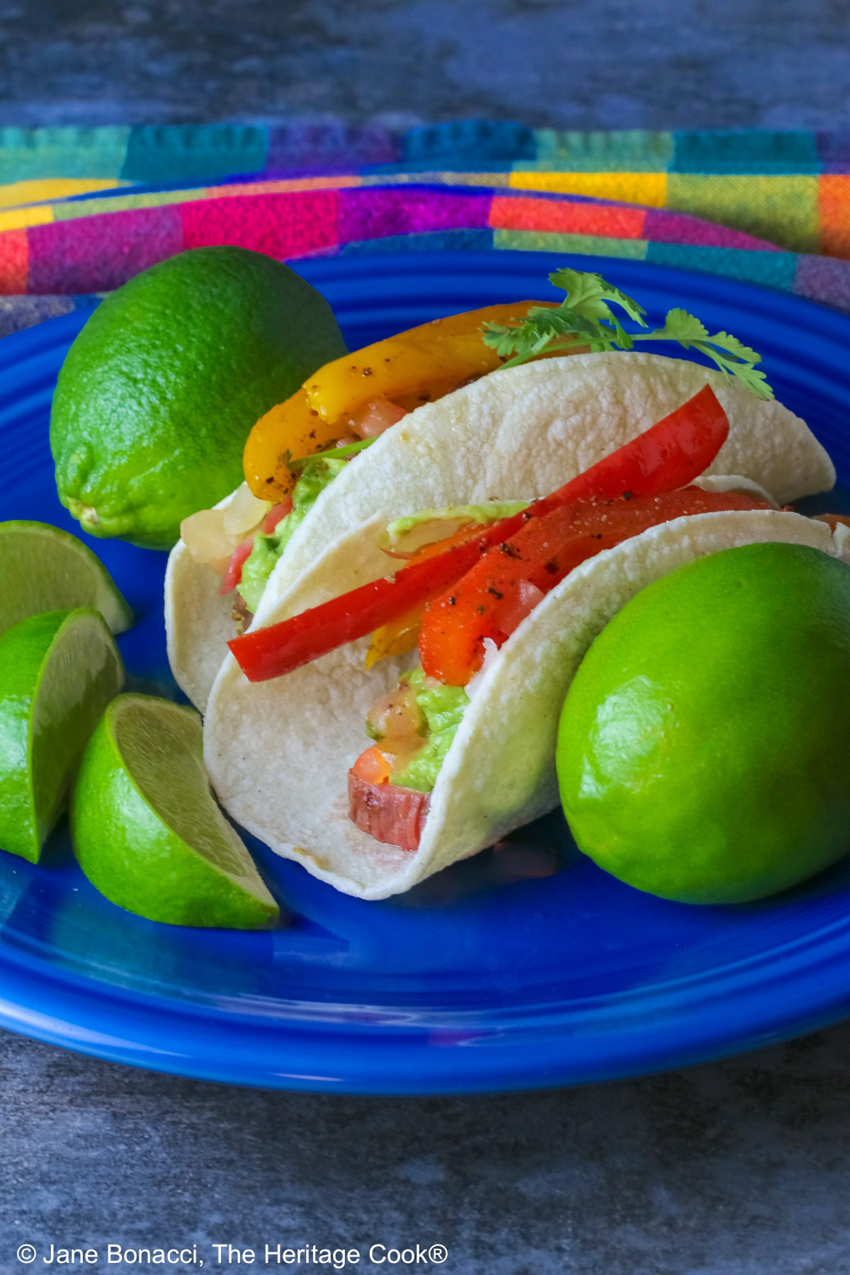 Two One-Pan Steak Fajitas tacos in corn tortillas on a dark blue plate, topped with guacamole, pico de gallo salsa, roasted onions, and bell pepper strips, surrounded by whole limes and lime wedges, with a multi-colored cloth behind and gray background © 2026 Jane Bonacci, The Heritage Cook.