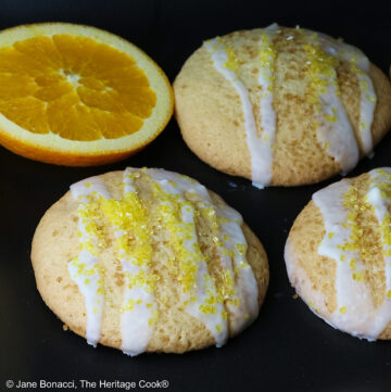 5 Large Orange and White Chocolate Sugar Cookies with two orange slices on a black rectangular platter with a blue checked cloth behind, each cookie drizzled with white chocolate glaze and sprinkled with yellow sparkling sugar © 2026 Jane Bonacci, The Heritage Cook.