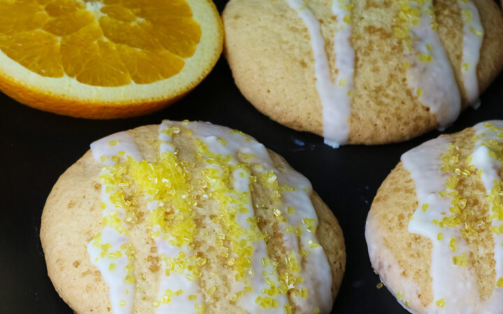 5 Large Orange and White Chocolate Sugar Cookies with two orange slices on a black rectangular platter with a blue checked cloth behind, each cookie drizzled with white chocolate glaze and sprinkled with yellow sparkling sugar © 2026 Jane Bonacci, The Heritage Cook.