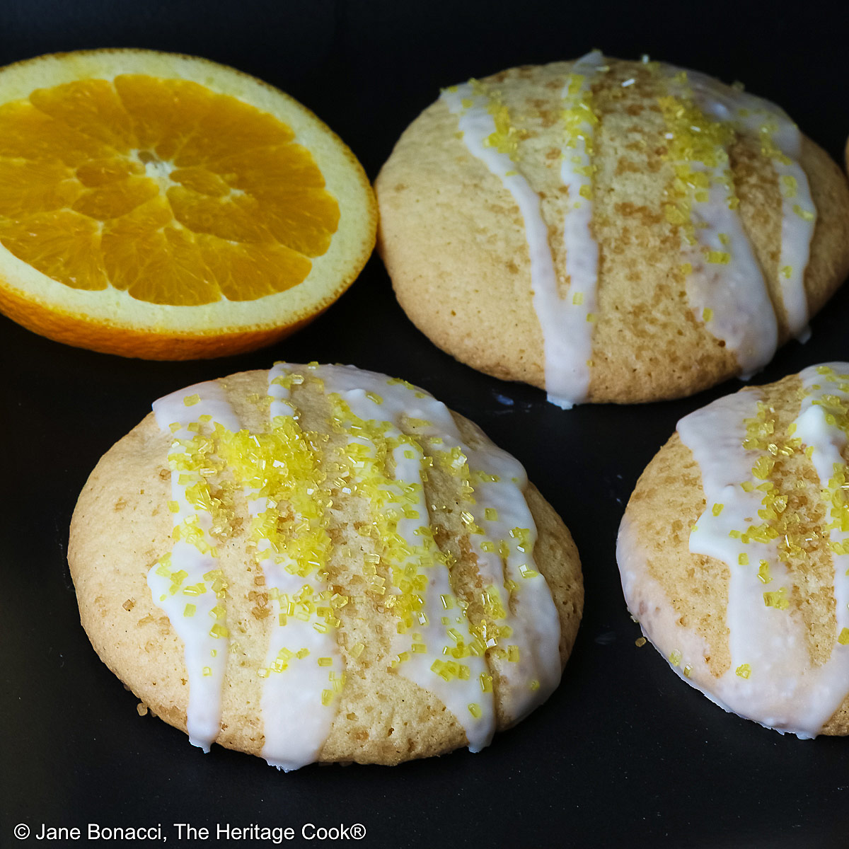 5 Large Orange and White Chocolate Sugar Cookies with two orange slices on a black rectangular platter with a blue checked cloth behind, each cookie drizzled with white chocolate glaze and sprinkled with yellow sparkling sugar © 2026 Jane Bonacci, The Heritage Cook.