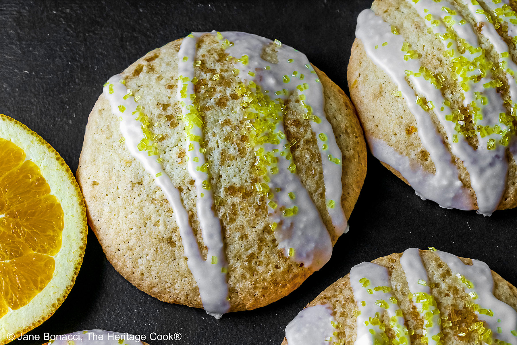 5 Large Orange and White Chocolate Sugar Cookies with two orange slices on a black rectangular platter with a blue checked cloth behind, each cookie drizzled with white chocolate glaze and sprinkled with yellow sparkling sugar © 2026 Jane Bonacci, The Heritage Cook.
