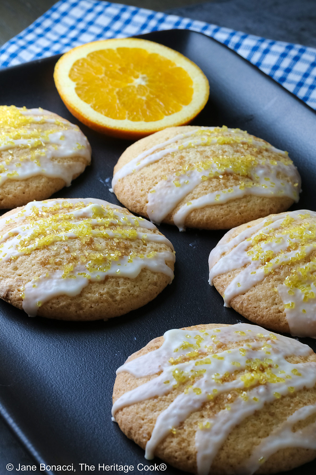 5 Large Orange and White Chocolate Sugar Cookies with two orange slices on a black rectangular platter with a blue checked cloth behind, each cookie drizzled with white chocolate glaze and sprinkled with yellow sparkling sugar © 2026 Jane Bonacci, The Heritage Cook.