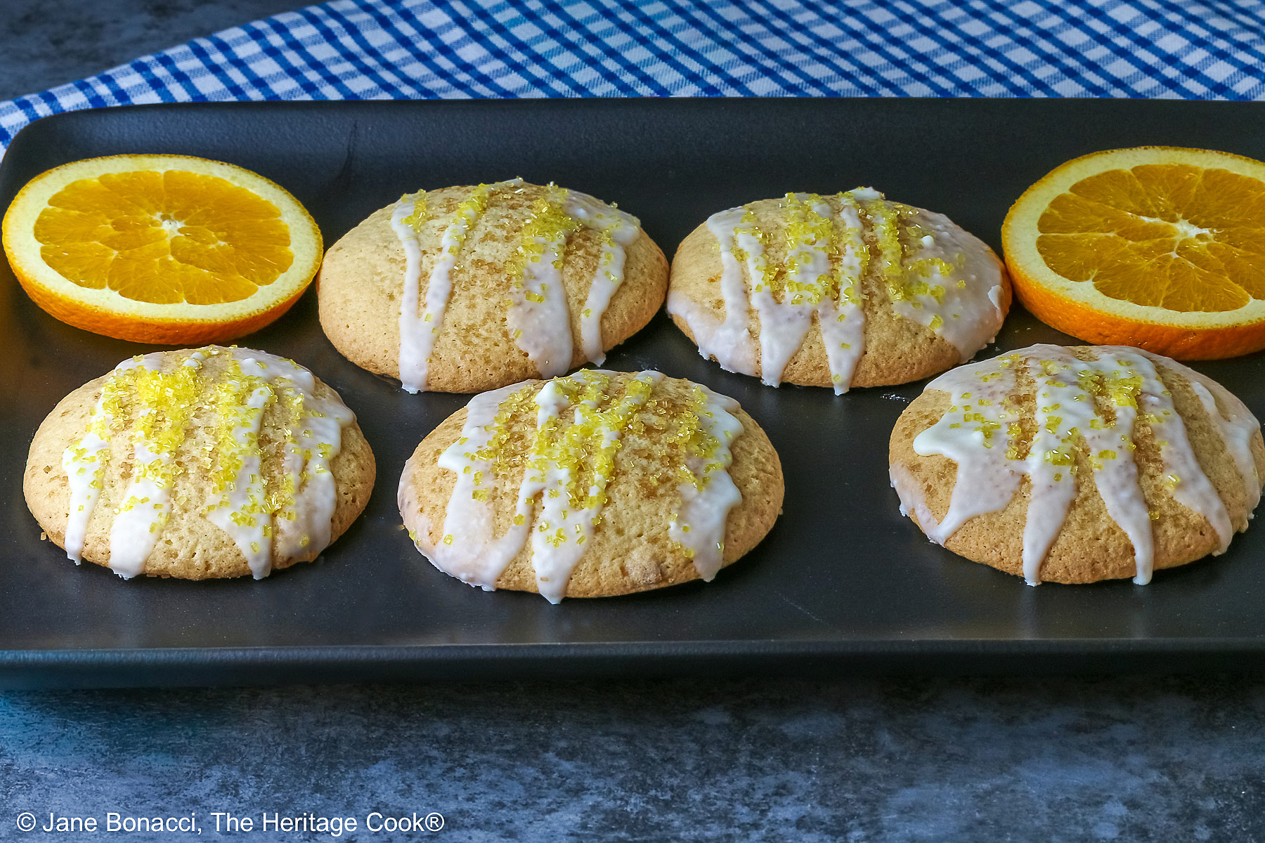 5 Large Orange and White Chocolate Sugar Cookies with two orange slices on a black rectangular platter with a blue checked cloth behind, each cookie drizzled with white chocolate glaze and sprinkled with yellow sparkling sugar © 2026 Jane Bonacci, The Heritage Cook.