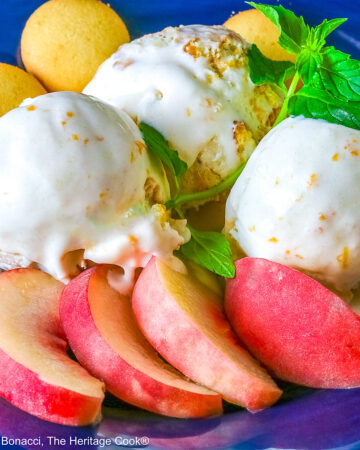 Three scoops of Peach Cobbler Ice Cream in a dark blue bowl with a broad rim, sitting on a deep pink napkin, with a sprig of mint, four slices of fresh peaches, some vanilla wafer cookies, and a whole peach in the foreground and a bottle of bourbon in the back © 2026 Jane Bonacci, The Heritage Cook.