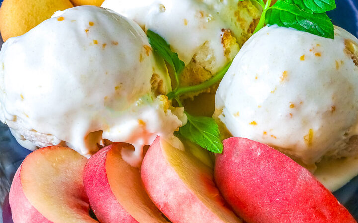 Three scoops of Peach Cobbler Ice Cream in a dark blue bowl with a broad rim, sitting on a deep pink napkin, with a sprig of mint, four slices of fresh peaches, some vanilla wafer cookies, and a whole peach in the foreground and a bottle of bourbon in the back © 2026 Jane Bonacci, The Heritage Cook.