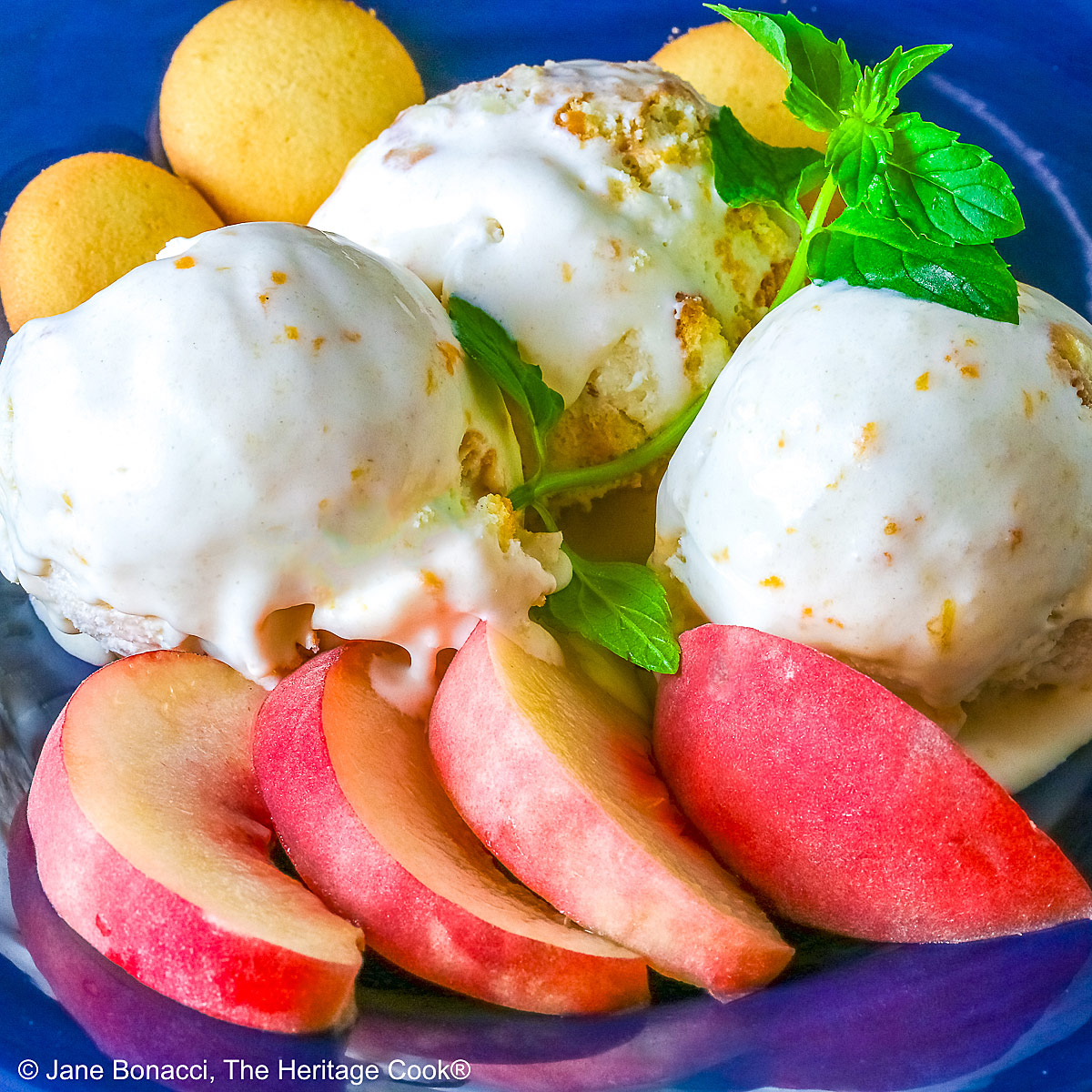 Three scoops of Peach Cobbler Ice Cream in a dark blue bowl with a broad rim, sitting on a deep pink napkin, with a sprig of mint, four slices of fresh peaches, some vanilla wafer cookies, and a whole peach in the foreground and a bottle of bourbon in the back © 2026 Jane Bonacci, The Heritage Cook.