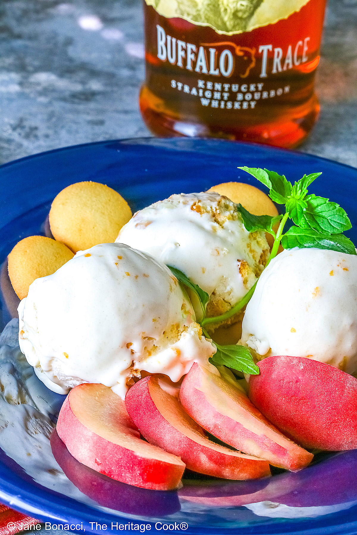 Three scoops of Peach Cobbler Ice Cream in a dark blue bowl with a broad rim, sitting on a deep pink napkin, with a sprig of mint, four slices of fresh peaches, some vanilla wafer cookies, and a whole peach in the foreground and a bottle of bourbon in the back © 2026 Jane Bonacci, The Heritage Cook.