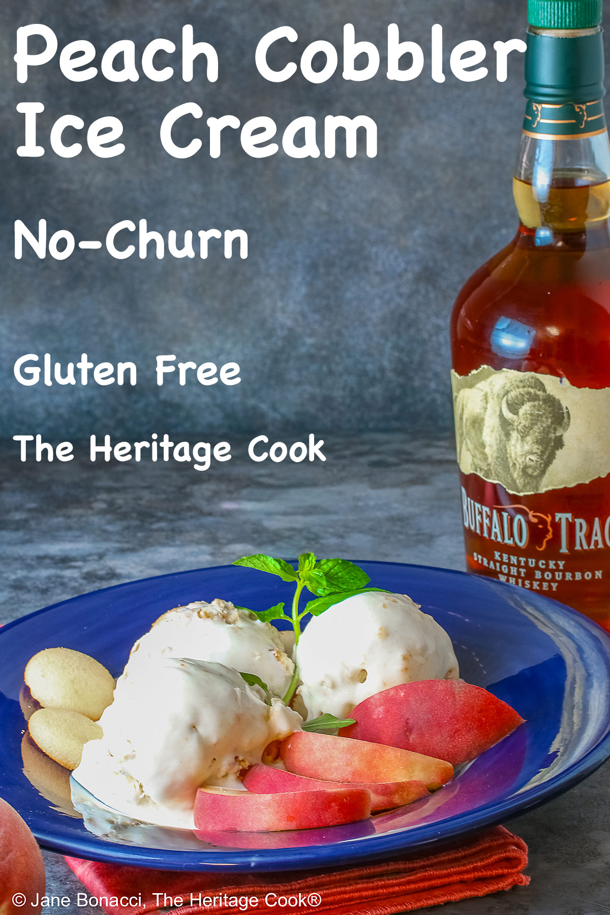 Three scoops of Peach Cobbler Ice Cream in a dark blue bowl with a broad rim, sitting on a deep pink napkin, with a sprig of mint, four slices of fresh peaches, some vanilla wafer cookies, and a whole peach in the foreground and a bottle of bourbon in the back © 2026 Jane Bonacci, The Heritage Cook.
