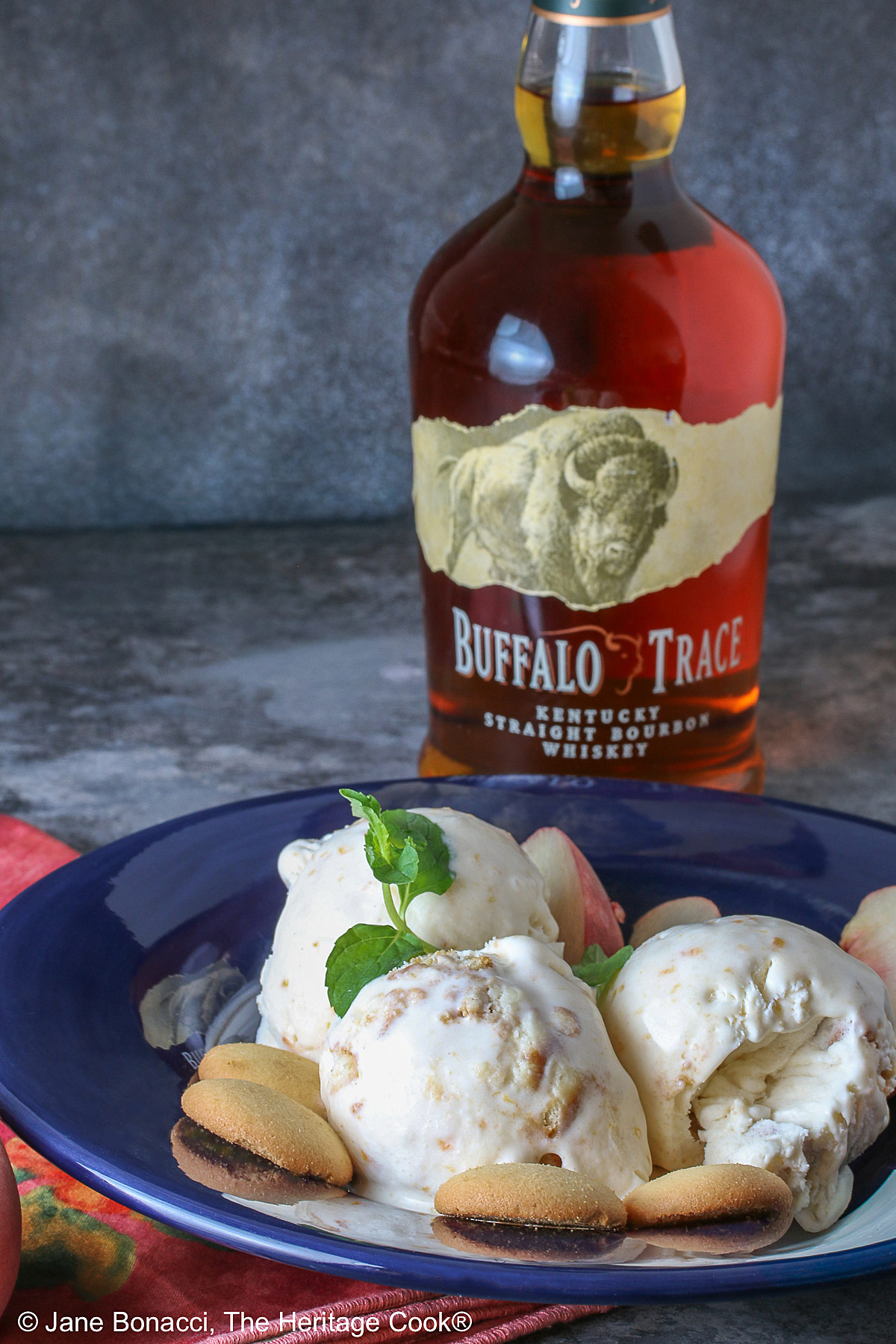Three scoops of Peach Cobbler Ice Cream in a dark blue bowl with a broad rim, sitting on a deep pink napkin, with a sprig of mint, four slices of fresh peaches, some vanilla wafer cookies, and a whole peach in the foreground and a bottle of bourbon in the back © 2026 Jane Bonacci, The Heritage Cook.