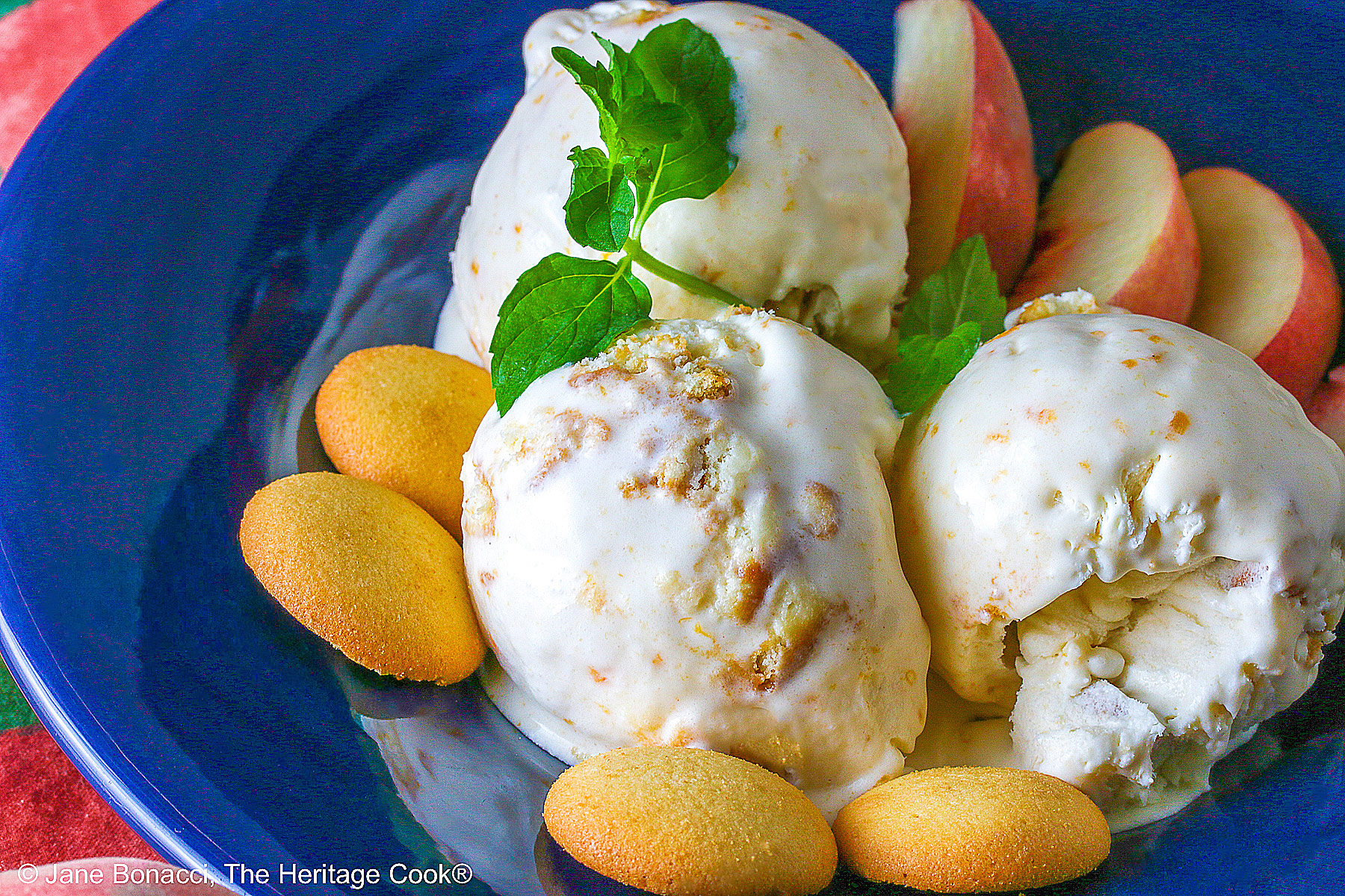 Three scoops of Peach Cobbler Ice Cream in a dark blue bowl with a broad rim, sitting on a deep pink napkin, with a sprig of mint, four slices of fresh peaches, some vanilla wafer cookies, and a whole peach in the foreground and a bottle of bourbon in the back © 2026 Jane Bonacci, The Heritage Cook.