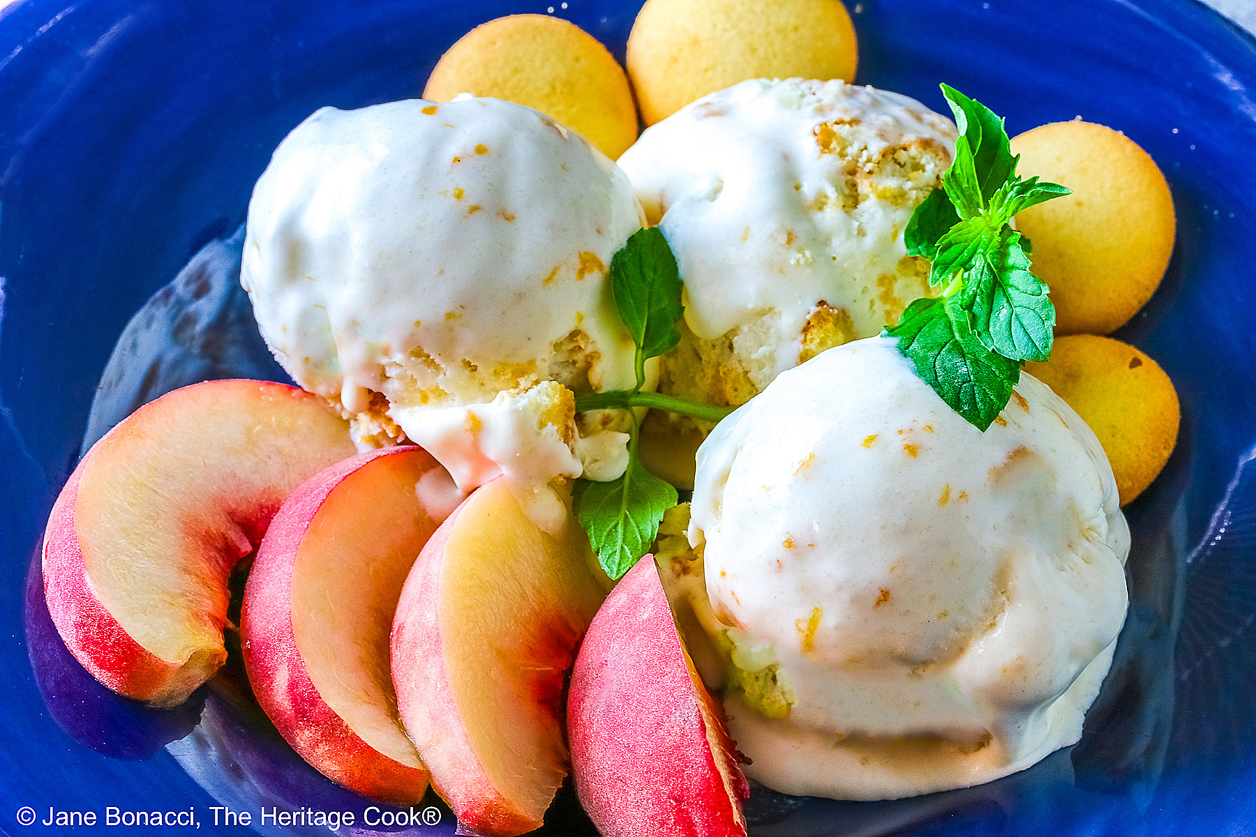 Three scoops of Peach Cobbler Ice Cream in a dark blue bowl with a broad rim, sitting on a deep pink napkin, with a sprig of mint, four slices of fresh peaches, some vanilla wafer cookies, and a whole peach in the foreground and a bottle of bourbon in the back © 2026 Jane Bonacci, The Heritage Cook.