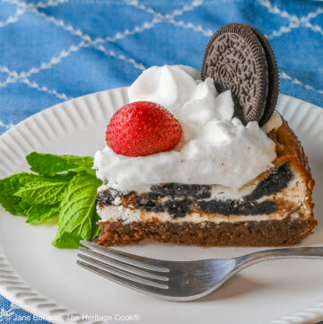 A slice of Scrumptious Oreo Brownie Cheesecake, topped with freshly made whipped cream, a whole Oreo cookie, and a whole strawberry, with a sprig of mint alongside © 2026 Jane Bonacci, The Heritage Cook.