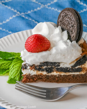 A slice of Scrumptious Oreo Brownie Cheesecake, topped with freshly made whipped cream, a whole Oreo cookie, and a whole strawberry, with a sprig of mint alongside © 2026 Jane Bonacci, The Heritage Cook.