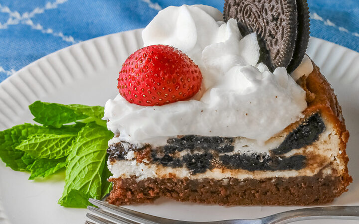 A slice of Scrumptious Oreo Brownie Cheesecake, topped with freshly made whipped cream, a whole Oreo cookie, and a whole strawberry, with a sprig of mint alongside © 2026 Jane Bonacci, The Heritage Cook.