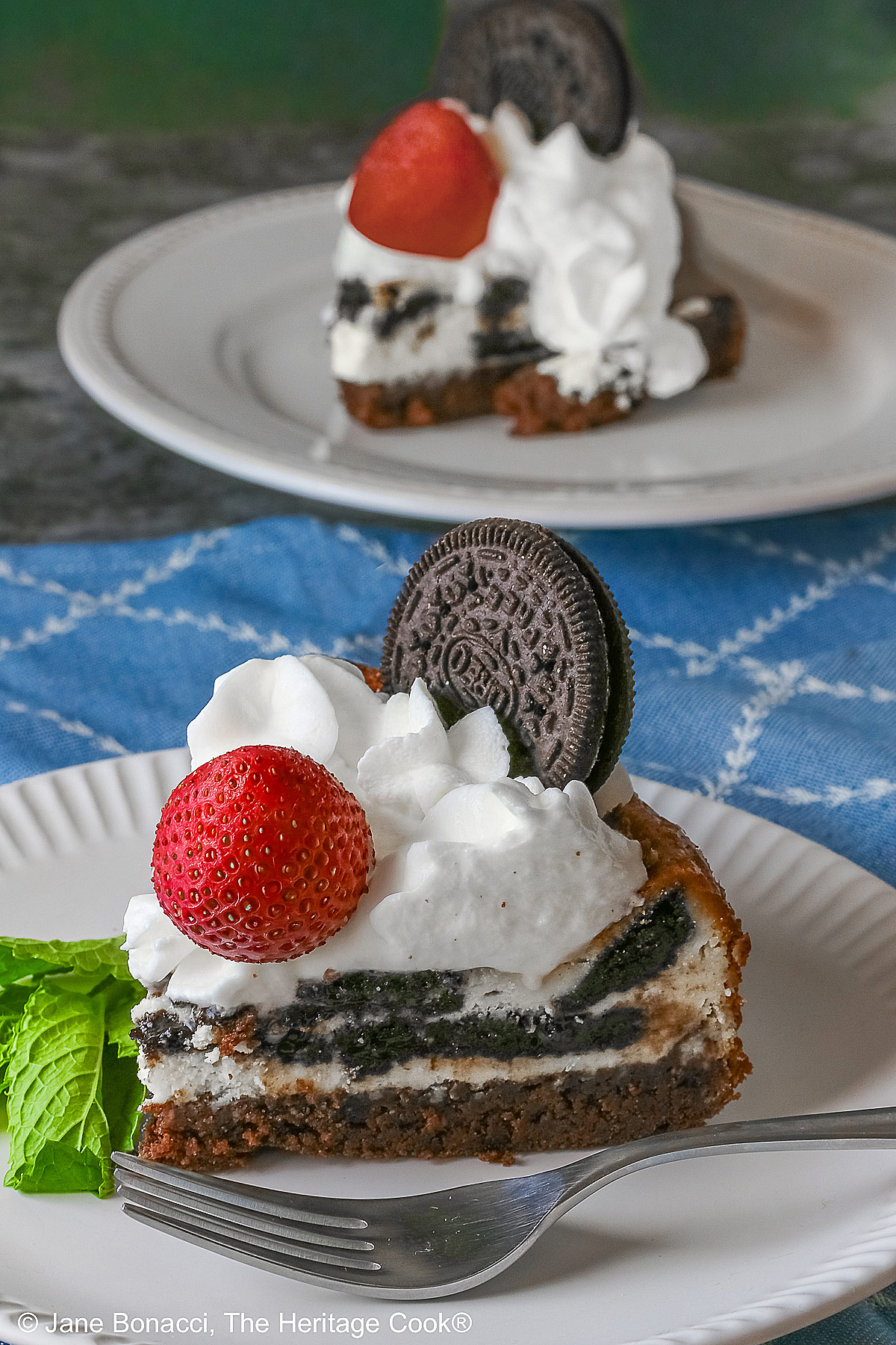 two plates, one behind the other, each with a slice of cheesecke; A slice of Scrumptious Oreo Brownie Cheesecake, topped with freshly made whipped cream, a whole Oreo cookie, and a whole strawberry, with a sprig of mint alongside © 2026 Jane Bonacci, The Heritage Cook.