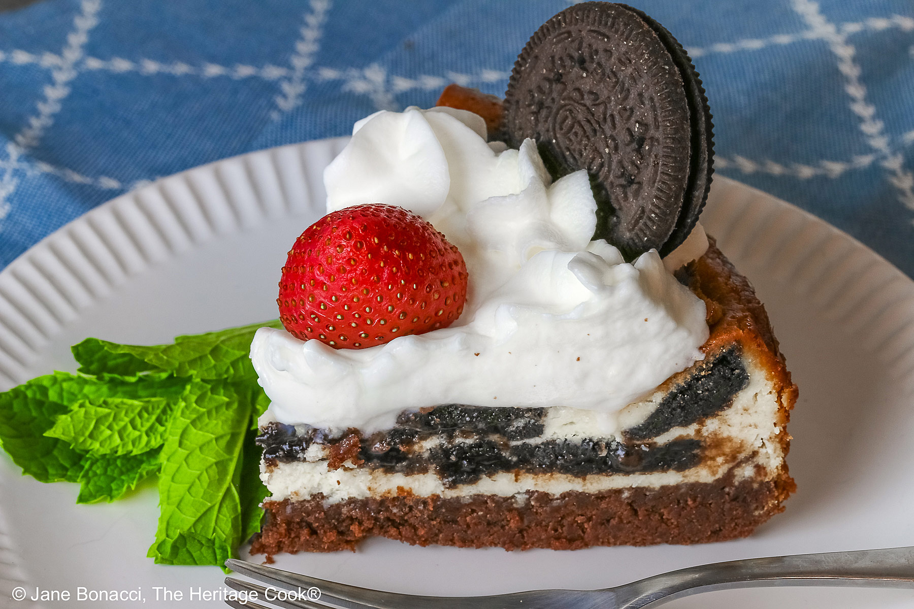 A slice of Scrumptious Oreo Brownie Cheesecake, topped with freshly made whipped cream, a whole Oreo cookie, and a whole strawberry, with a sprig of mint alongside © 2026 Jane Bonacci, The Heritage Cook.