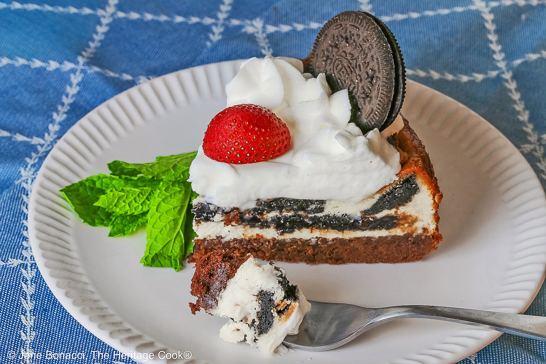 Bite on a fork in front of a slice of cheesecake with mint sprig, strawberry, and whole Oreo cookie.