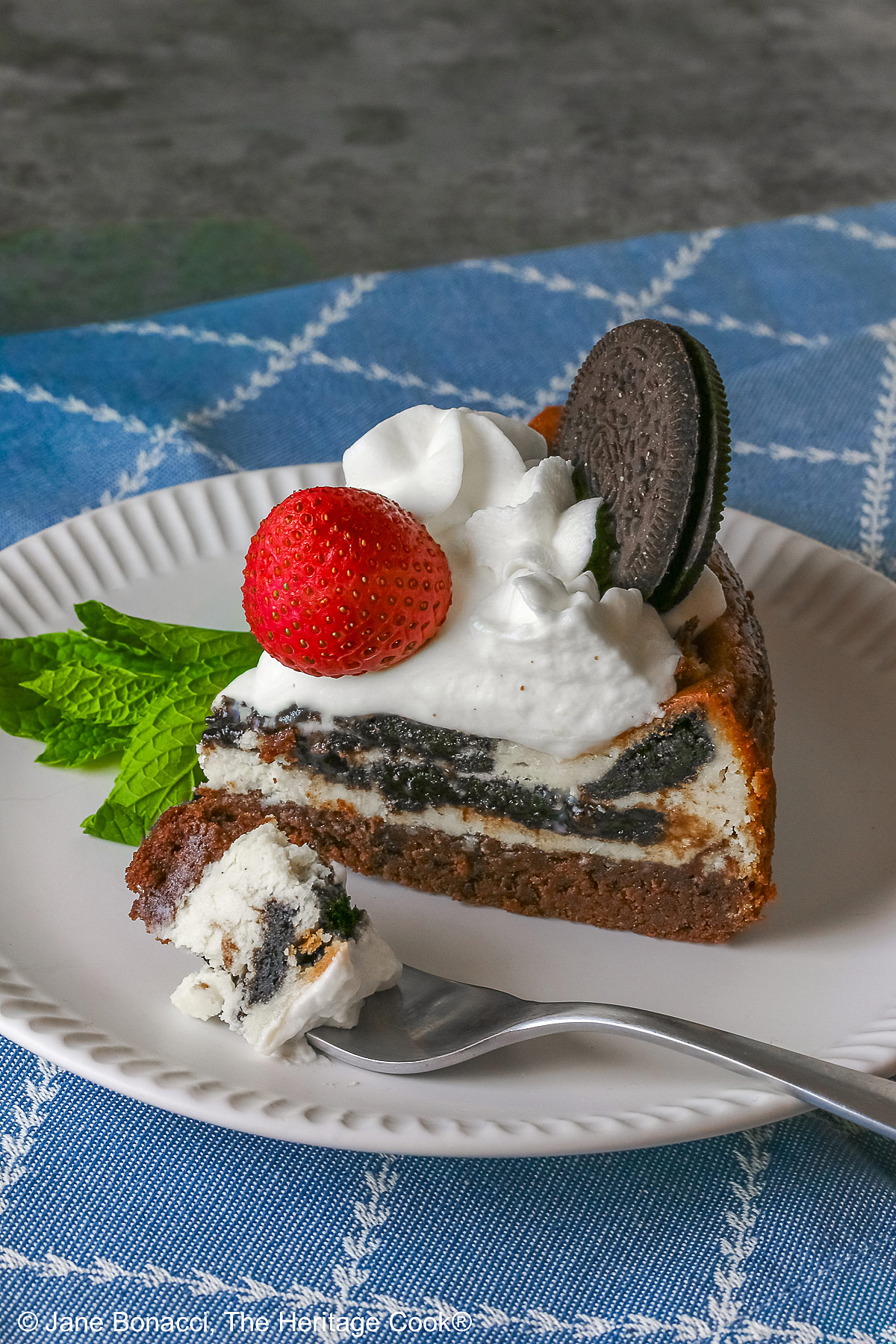 A slice of Scrumptious Oreo Brownie Cheesecake, topped with freshly made whipped cream, a whole Oreo cookie, and a whole strawberry, with a sprig of mint alongside © 2026 Jane Bonacci, The Heritage Cook.